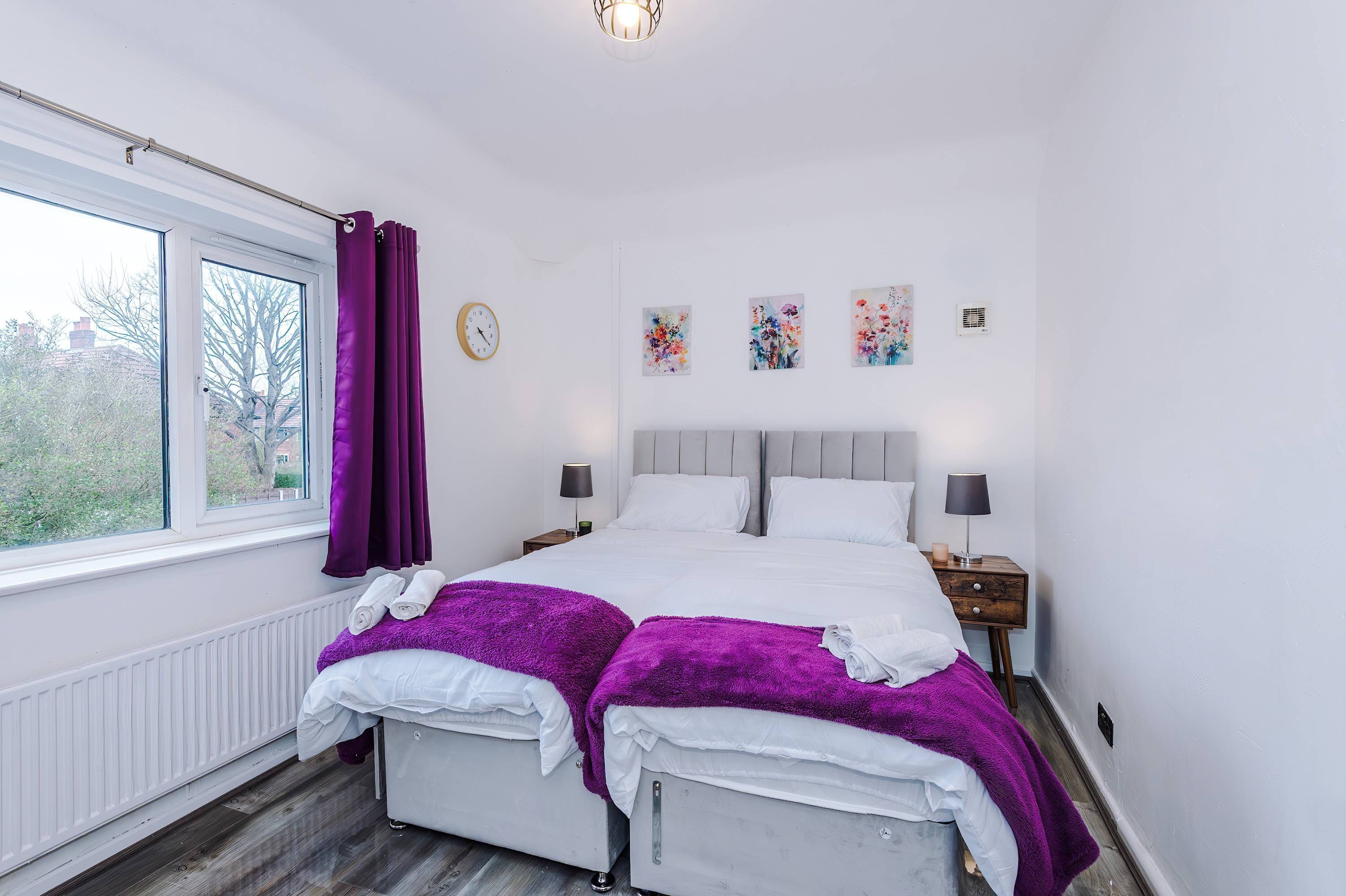 Une chambre à coucher comprenant un lit, deux tables de chevet, une fenêtre avec des rideaux, un radiateur et des œuvres d’art murales.