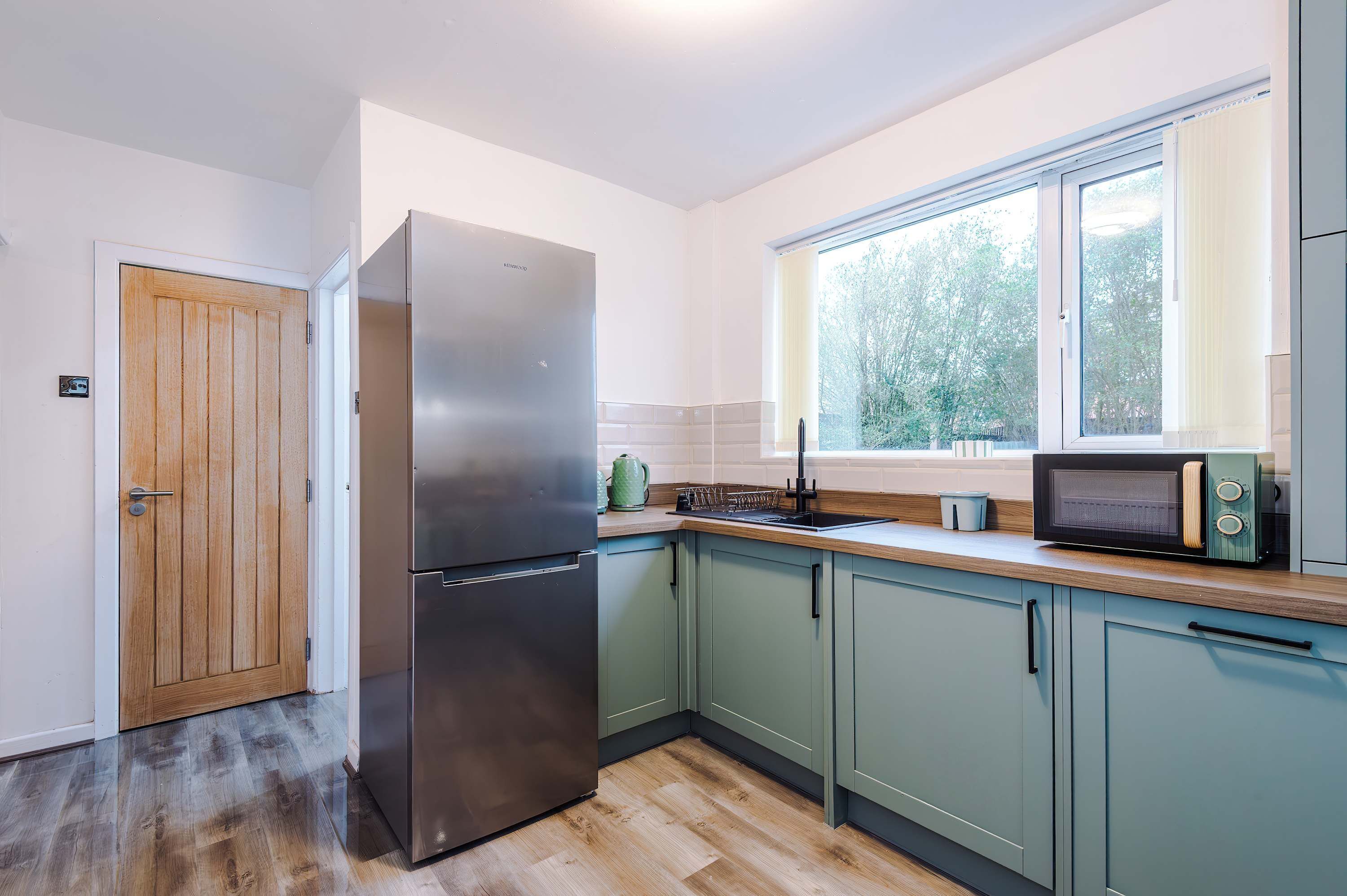A modern kitchen with a stainless steel refrigerator, a microwave, and a window with a view of trees.