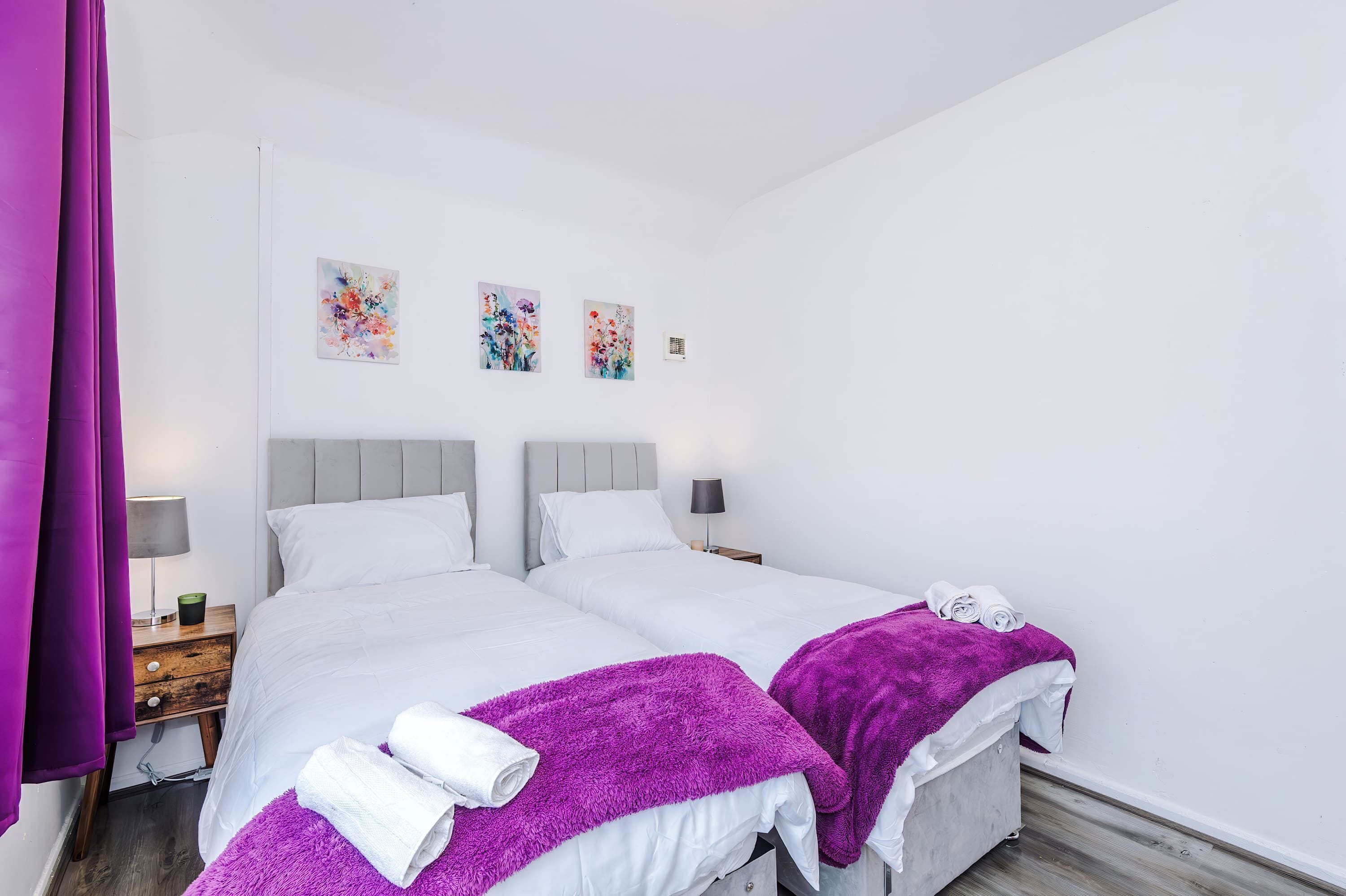 A double room with two beds, white bedding, purple throw blankets, and artwork on the wall.