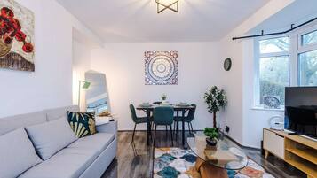 A modern living room with a grey sofa, a dining table with chairs, a wall-mounted artwork, a floor lamp, and a large window.