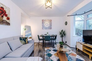 A modern living room with a grey sofa, a dining table with chairs, a wall-mounted artwork, a floor lamp, and a large window. - Large 3BR Near Airport Hospital Free Parking (Manchester)