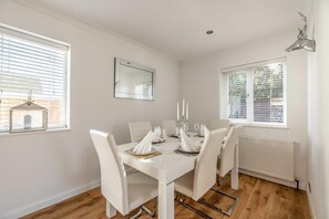 Un coin repas avec une table blanche dressée pour quatre personnes, des chaises blanches, un miroir au mur et une fenêtre avec des stores.