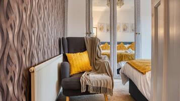 Un dormitorio con un sillón oscuro, un cojín amarillo y un espejo que refleja una cama con ropa de cama amarilla.