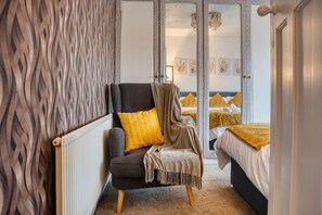 Un dormitorio con un sillón oscuro, un cojín amarillo y un espejo que refleja una cama con ropa de cama amarilla.