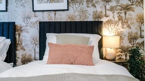 En säng med vita sängkläder och en rutig kudde, en mörk sänggavel och en vägg med ett blommigt tapetmönster.