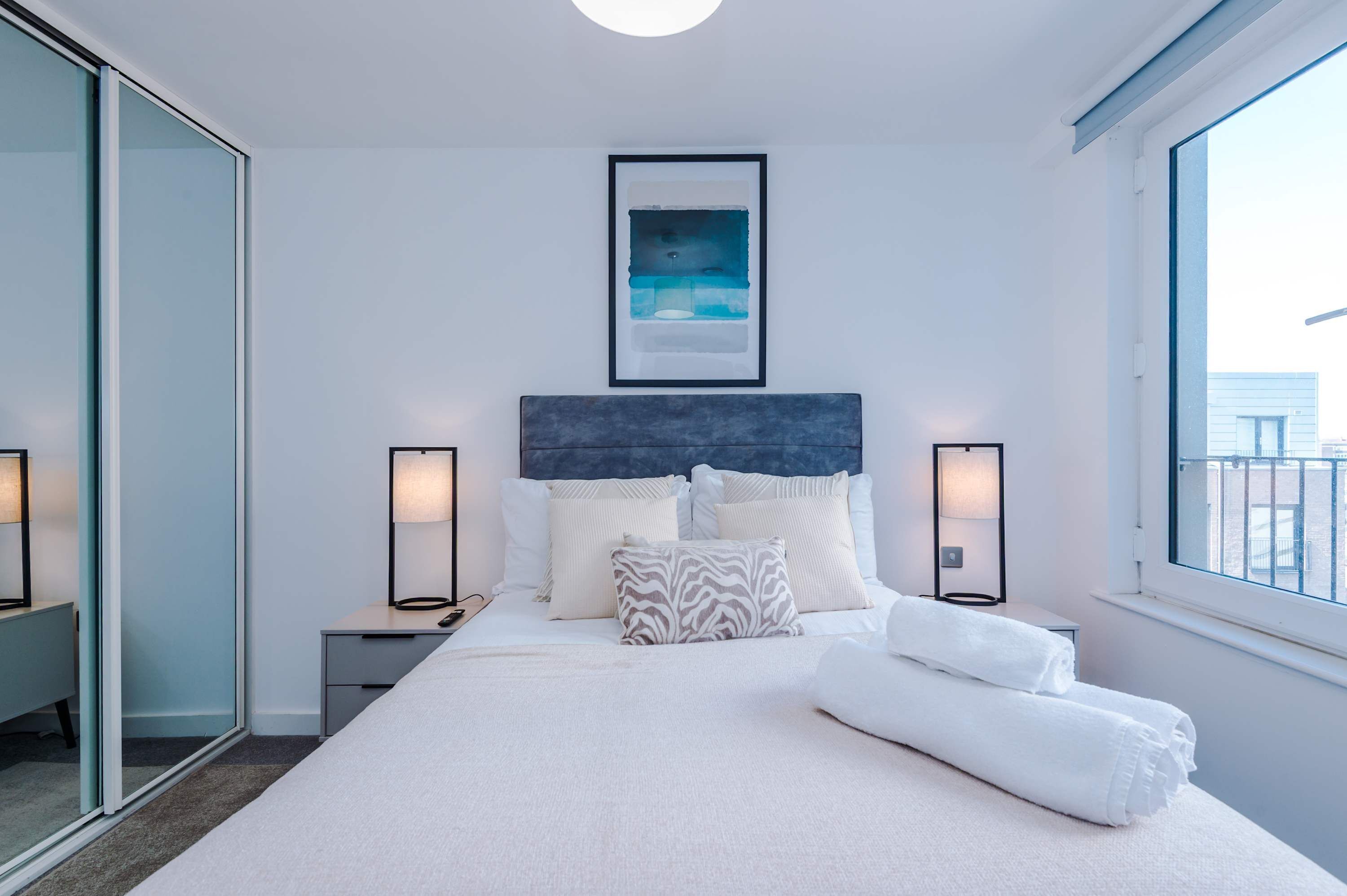 A hotel room with a large bed, a headboard, a framed picture above the bed, two bedside lamps, and a view through the window.