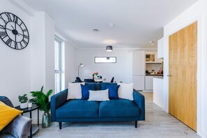 A modern living room with a blue sofa, a large wall clock, and a kitchen in the background. - Modern 2 Bed Duplex Free Parking Wi Fi (Salford)
