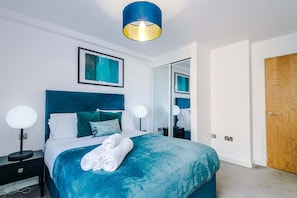 A modern hotel room with a large bed, a blue headboard, two bedside lamps, and a ceiling light fixture. - Modern 2 Bed Duplex Free Parking Wi Fi (Salford)