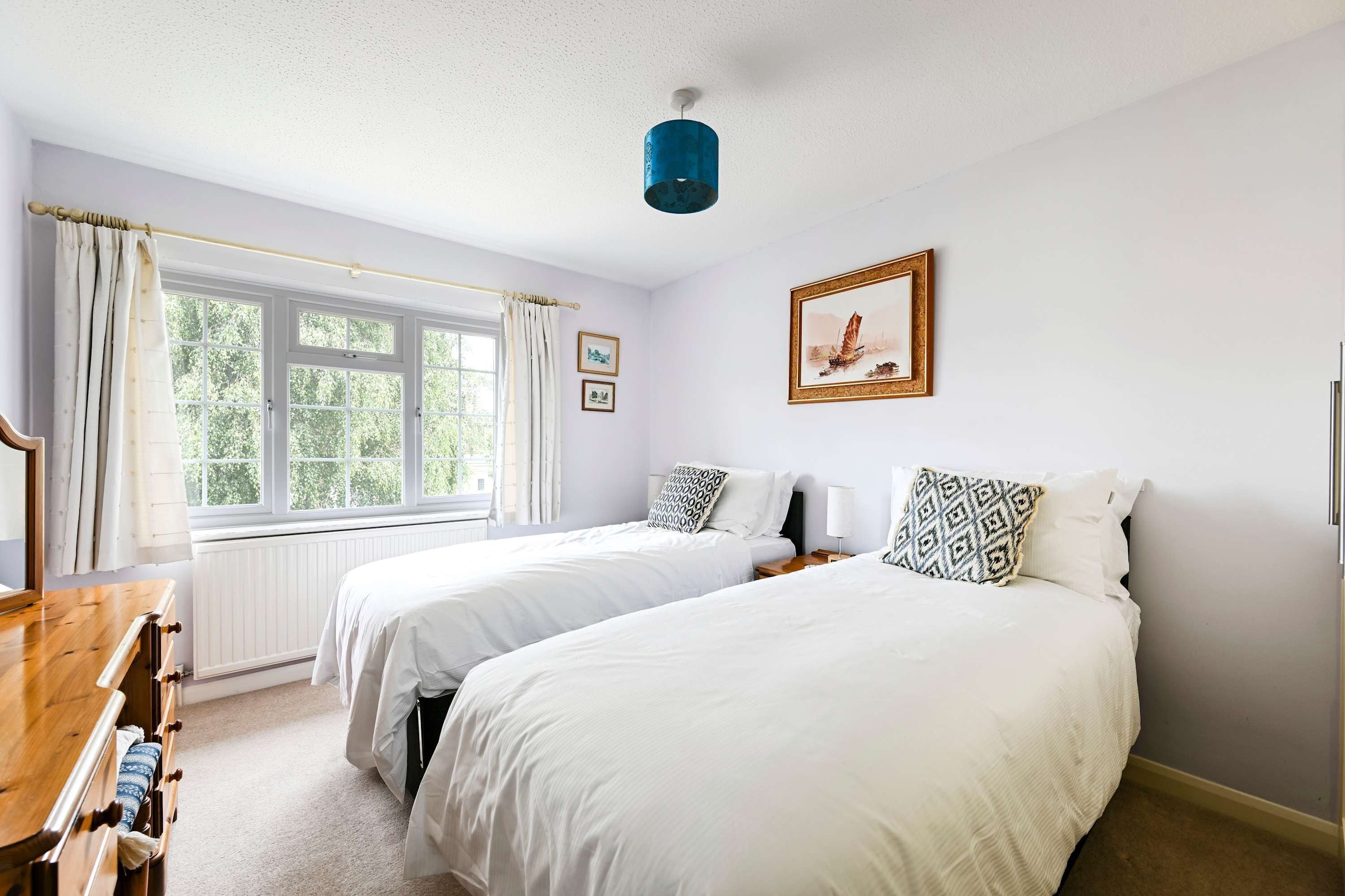 A bedroom with two beds, a window with curtains, a wooden dresser, and a framed picture on the wall.