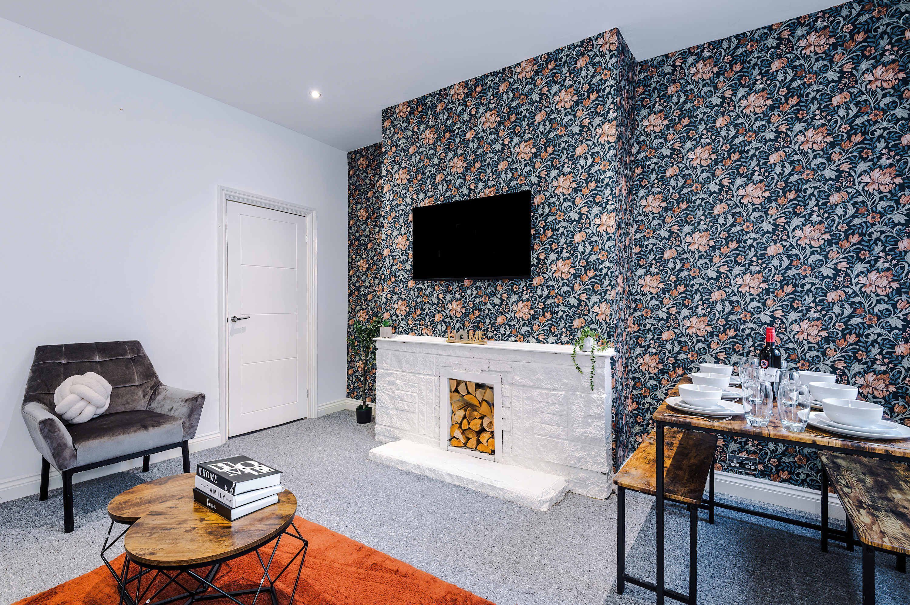 A living room with a fireplace, a flat-screen TV mounted on the wall, a dining table set with plates and glasses, and a comfortable armchair.