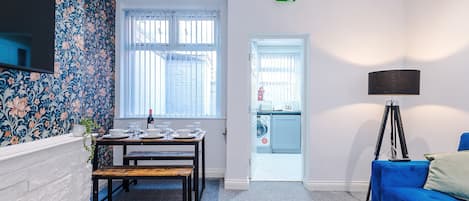 A modern living room with a blue sofa, a dining table set for four, and a kitchen area visible through an open door.
