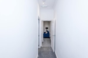 A narrow hallway with white walls and a grey carpet leading to a room with a blue bed and a black TV. - 3 Bedroom Town House - Etihad Stadium - Free Parking (Manchester)