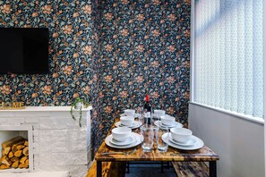 Un coin repas agrémenté d’un papier peint à motifs floraux, d’une cheminée et d’une fenêtre avec un store à motifs.