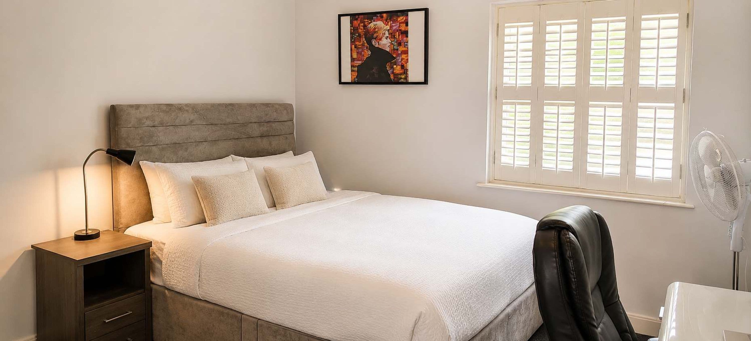 A bedroom with a bed, bedside lamp, a chair, a desk, a fan, and a window with blinds.