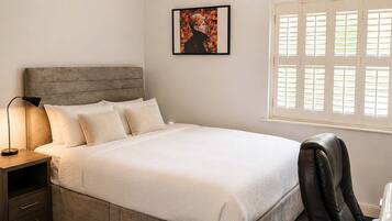 A bedroom with a bed, bedside lamp, a chair, a desk, a fan, and a window with blinds.