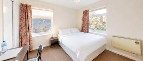 A bedroom with a bed, a desk with a chair, a radiator, and two windows with curtains.