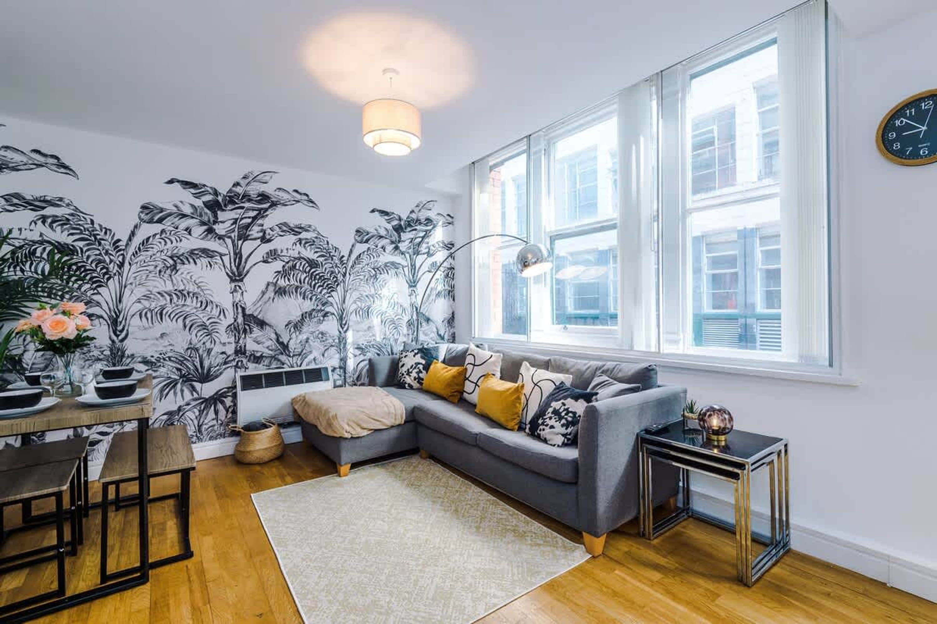 Een moderne woonkamer met een grijze bank, een eettafel met stoelen, een groot raam met uitzicht en een wand met een mural van een tropische plant.