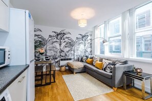 Ein modernes Wohnzimmer mit einem grauen Sofa, einem Esstisch mit Stühlen, einer Küchenzeile inklusive Mikrowelle und einem großen Fenster, das für natürliches Licht sorgt.
