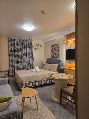 A hotel room with a bed, a round table, a chair, a TV, and a wall-mounted clock. - Condo near Mactan Beaches (Lapu-Lapu)