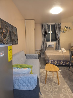 A compact room with a bed, a sofa, a small table, and a window with curtains. - Condo near Mactan Beaches (Lapu-Lapu)