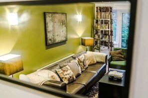 Ein Wohnzimmer mit grüner Wand, einer gepolsterten Couch mit gemusterten Kissen, einem Beistelltisch mit Büchern und einem Bücherregal mit Dekorationen.