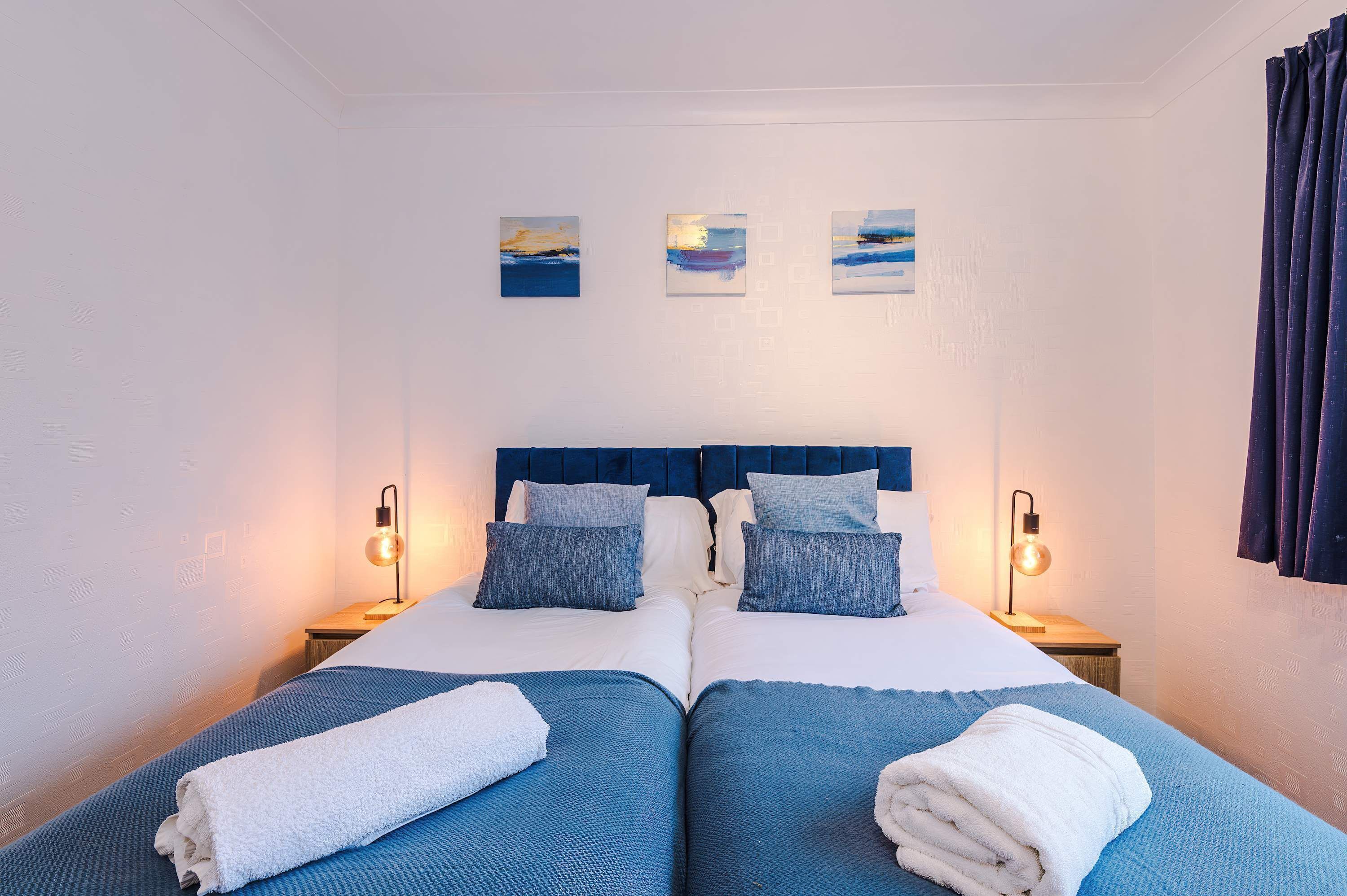 A double bed with a blue headboard, two bedside tables with lamps, and three wall-mounted paintings.