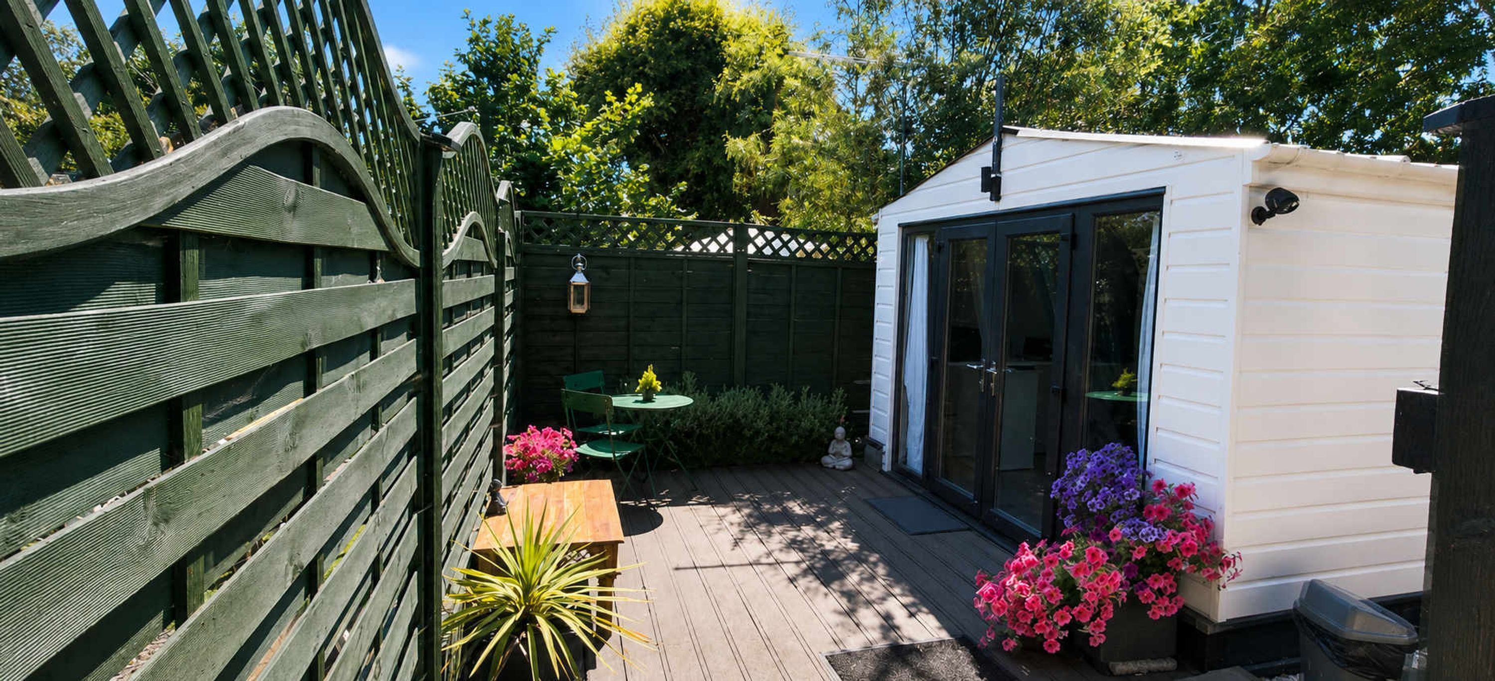 Une terrasse en bois avec une petite table et des chaises, un abri de jardin blanc et un espace aménagé en jardin avec des fleurs.