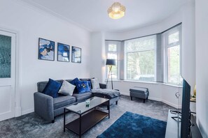 Ein modernes Wohnzimmer mit einem grauen Sofa, einem Couchtisch und einem blauen Teppich. An der Wand hängen drei gerahmte Kunstwerke, eine Stehlampe und ein großes Fenster mit Jalousien.