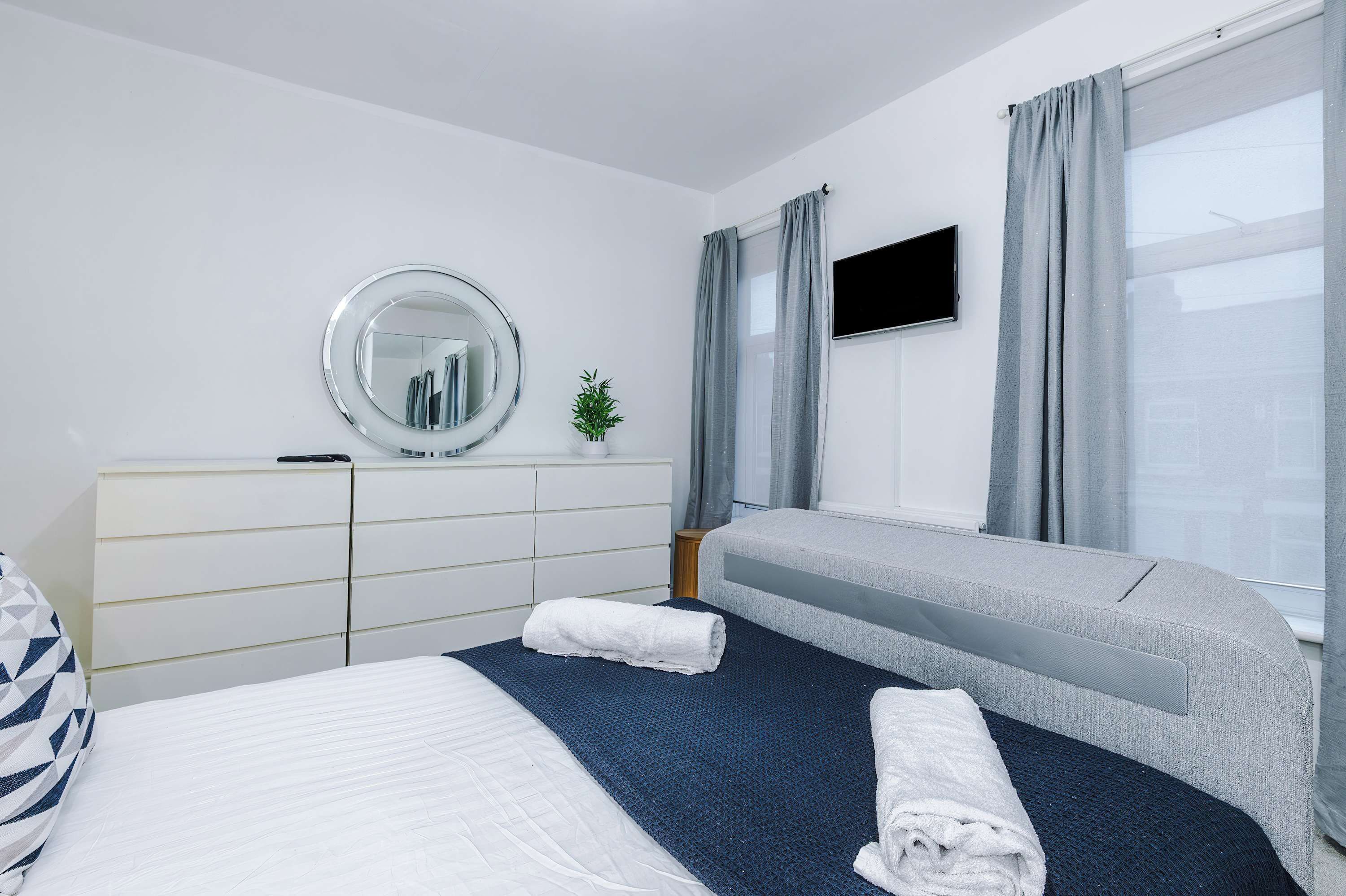 A hotel room with a bed, a bedside table, a mirror, a TV, and a window with curtains.