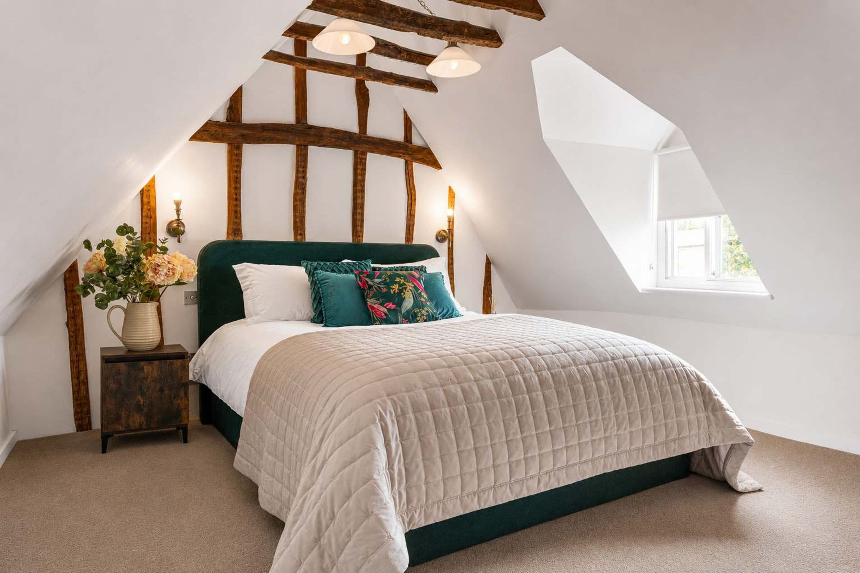 A bedroom with a sloped ceiling, a bed with a quilted duvet, a nightstand with a vase of flowers, and a window providing natural light.