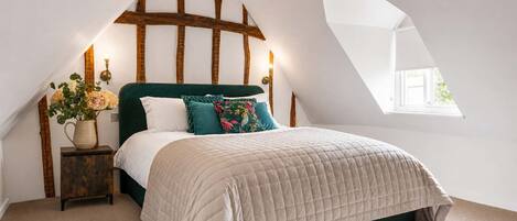 A bedroom with a sloped ceiling, a bed with a quilted duvet, a nightstand with a vase of flowers, and a window providing natural light.