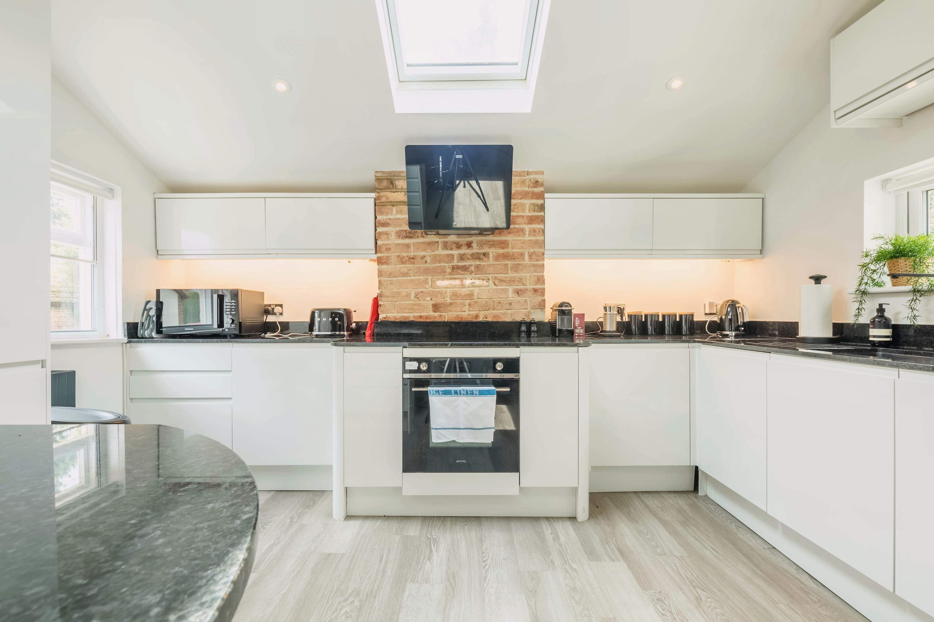 Una cocina moderna con muebles blancos, una pared de ladrillo decorativa, horno empotrado y tragaluz.