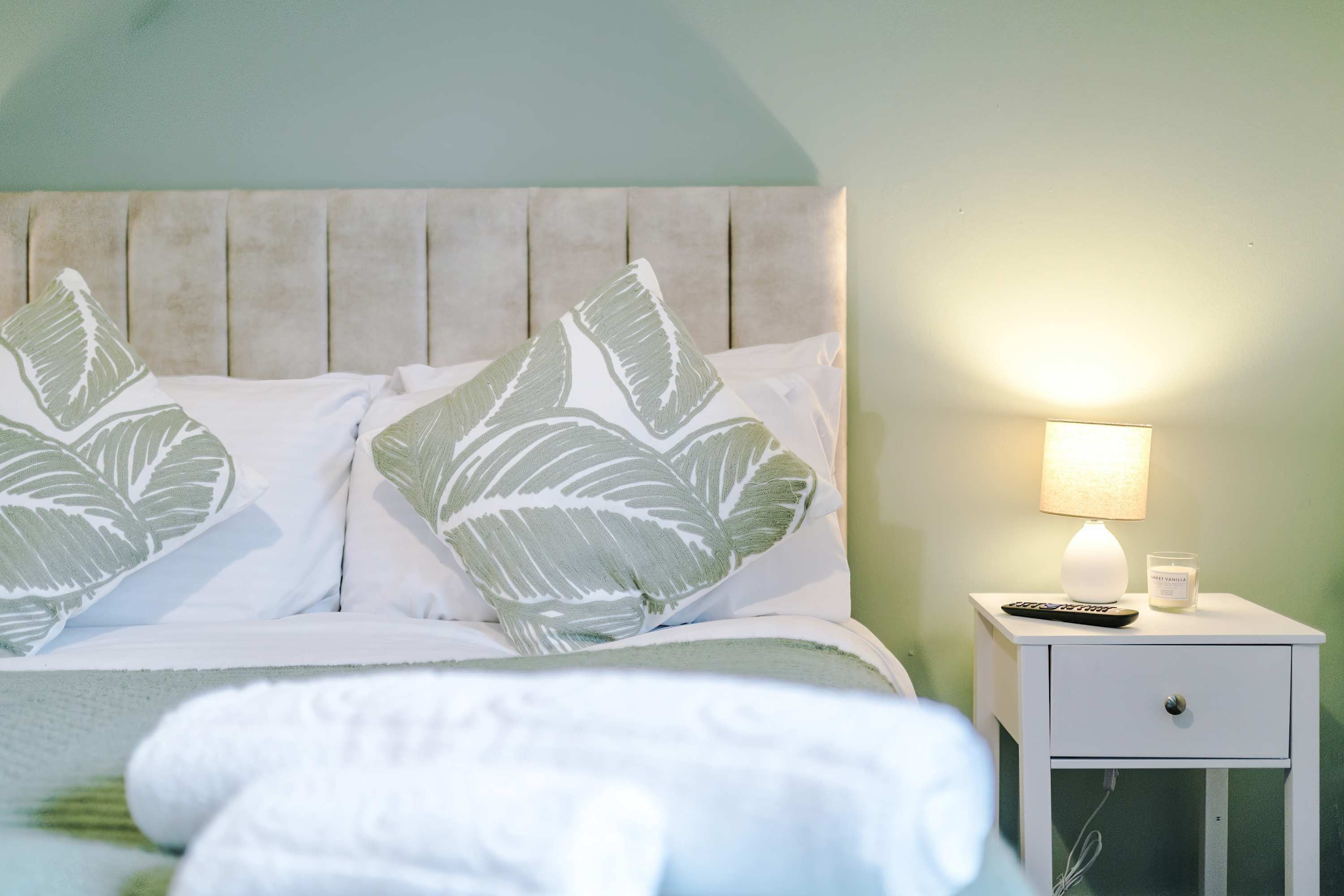 A bed with a leaf-patterned pillow, a bedside lamp, a remote control, and a candle on a bedside table.