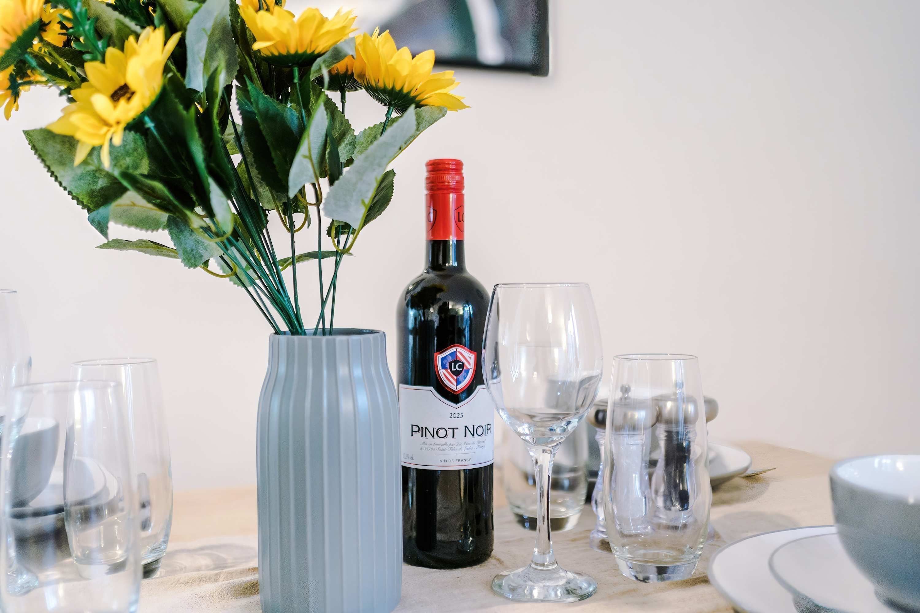 Ett dukat bord med en flaska Pinot Noir-vin, en vas med gula blommor och flera tomma vinglas.