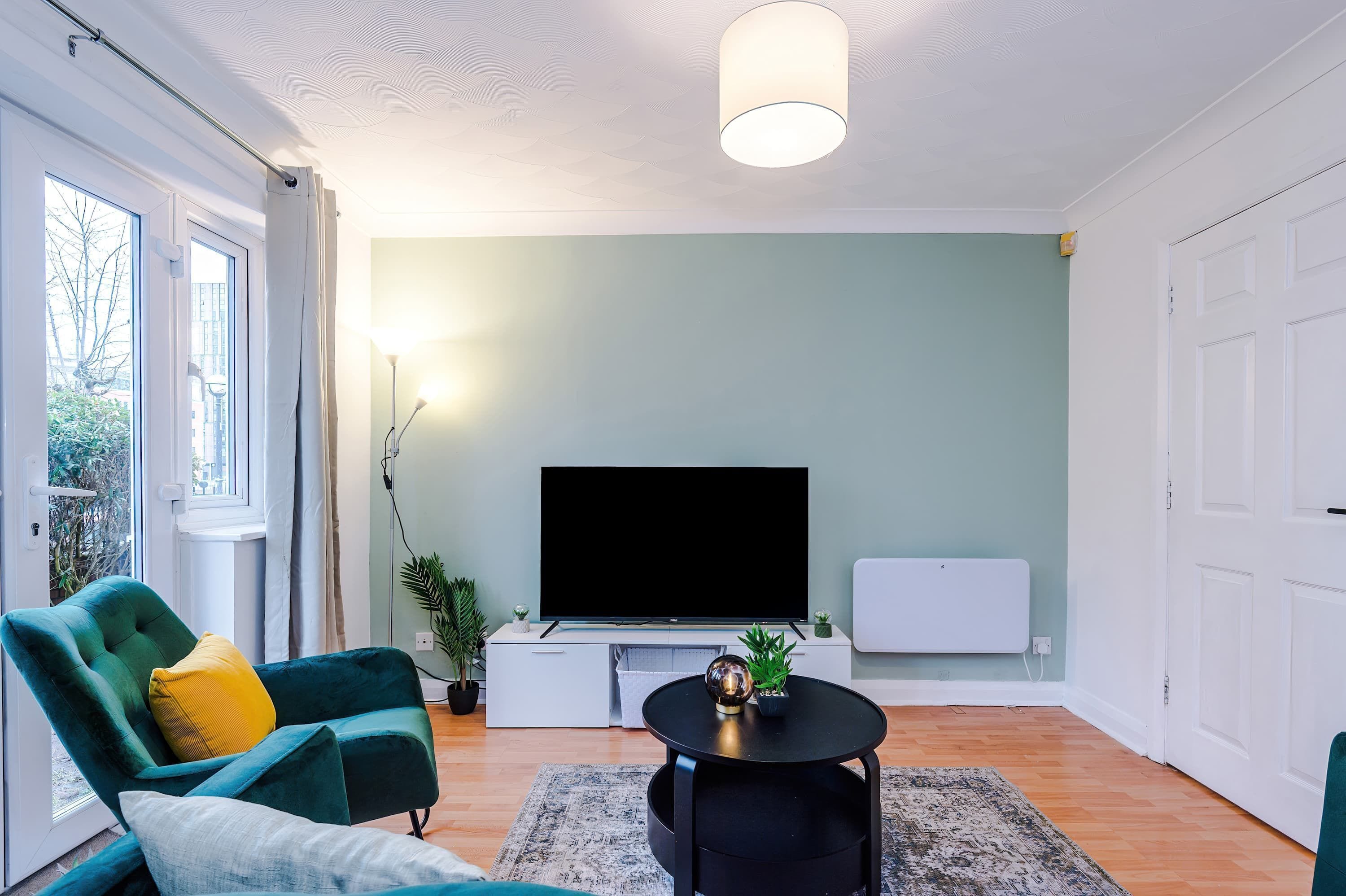 Una sala de estar moderna con un televisor de pantalla plana, un sillón verde azulado, una mesita redonda y un radiador.