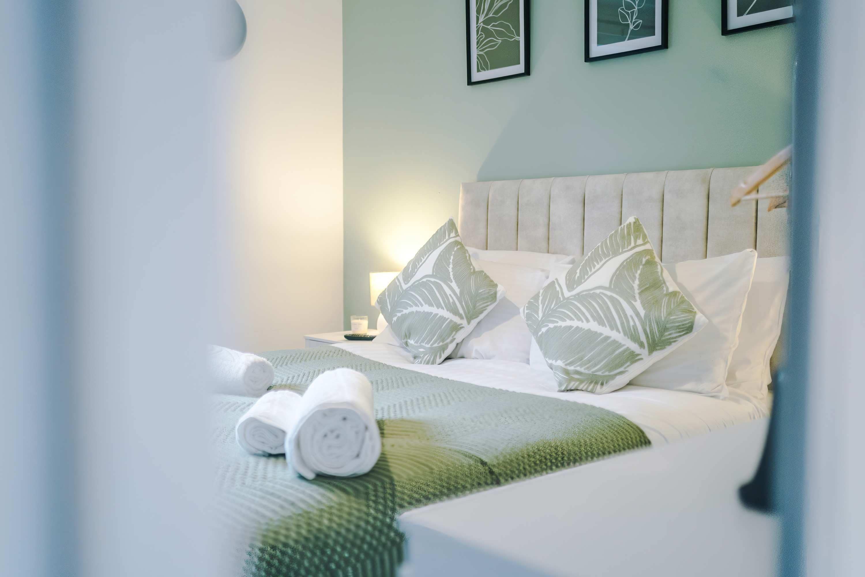 A neatly made bed with a green and white leaf-patterned bedspread, two pillows with similar leaf designs, and rolled white towels on the bed.