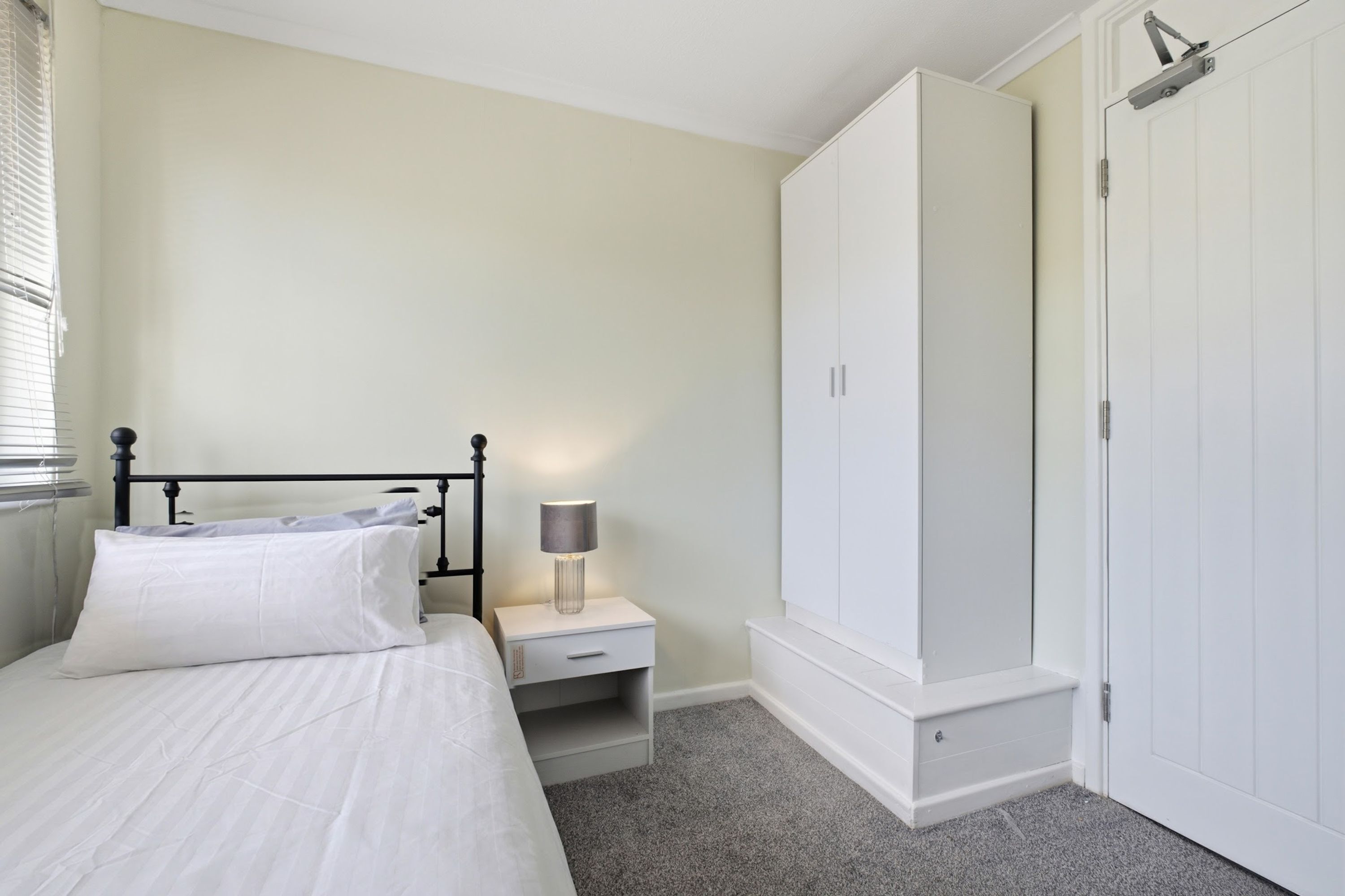 A bedroom with a bed, nightstand, lamp, wardrobe, and a door.
