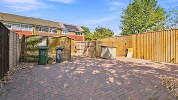 A paved backyard with a wooden fence, a brick pathway, and a house with a tiled roof.