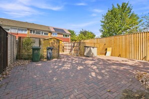 A paved backyard with a wooden fence, a brick pathway, and a house with a tiled roof. - Spacious 3-bed House in Waltham Cross Sleeps 6 (Waltham Cross)