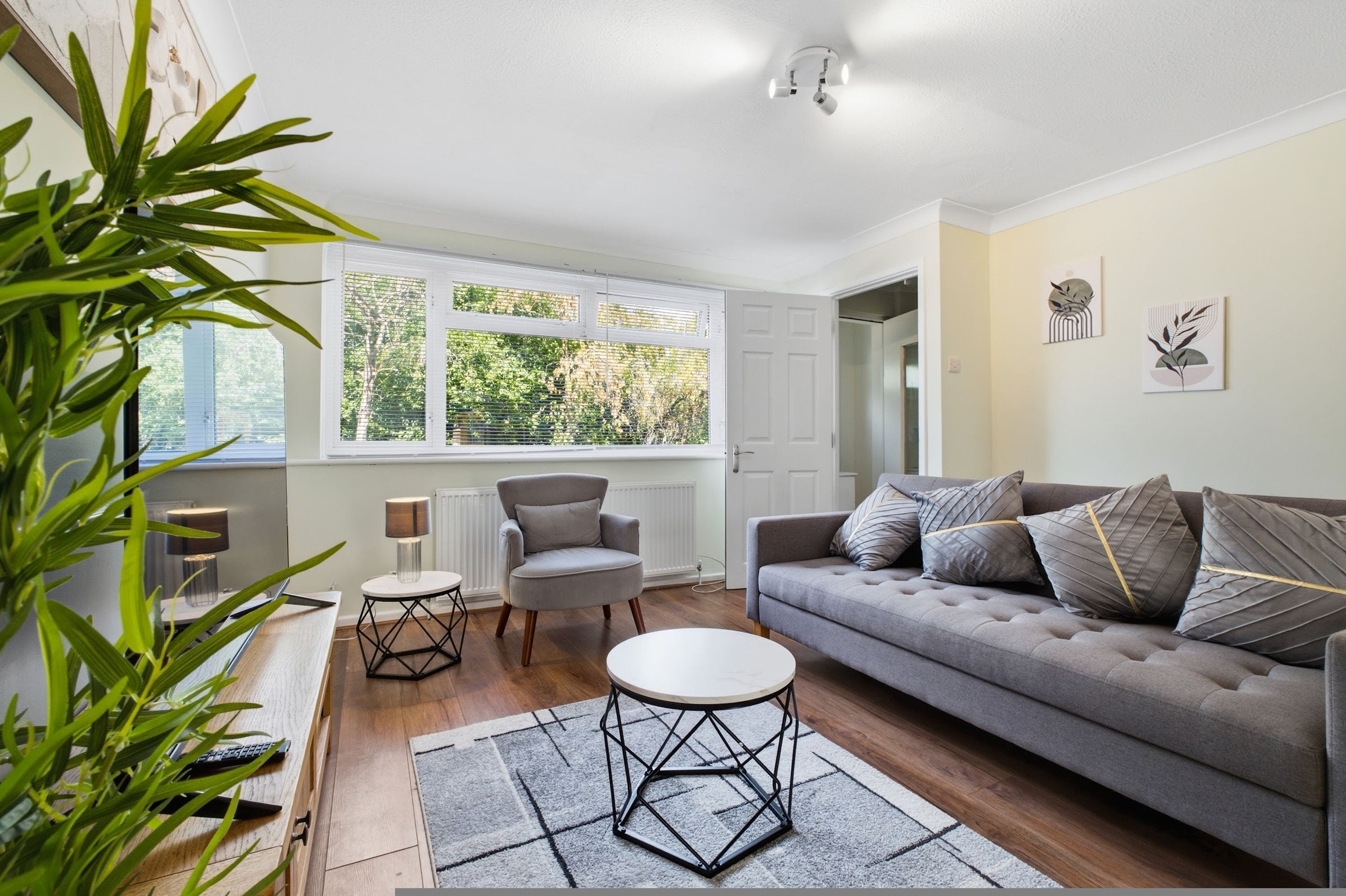 A modern living room with a grey sofa, a round coffee table, and a chair. There are two windows with blinds, a radiator, and a plant in the corner.