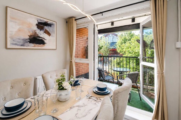 Un comedor con una mesa dispuesta para dos personas, un florero con flores y vista a un jardín a través de un ventanal.