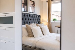 A bedroom with a tufted headboard, a bed with multiple pillows, a bedside table with a lamp, and a window with curtains. - Prime 3BR City Flat Balcony Walk to Shops (Manchester)