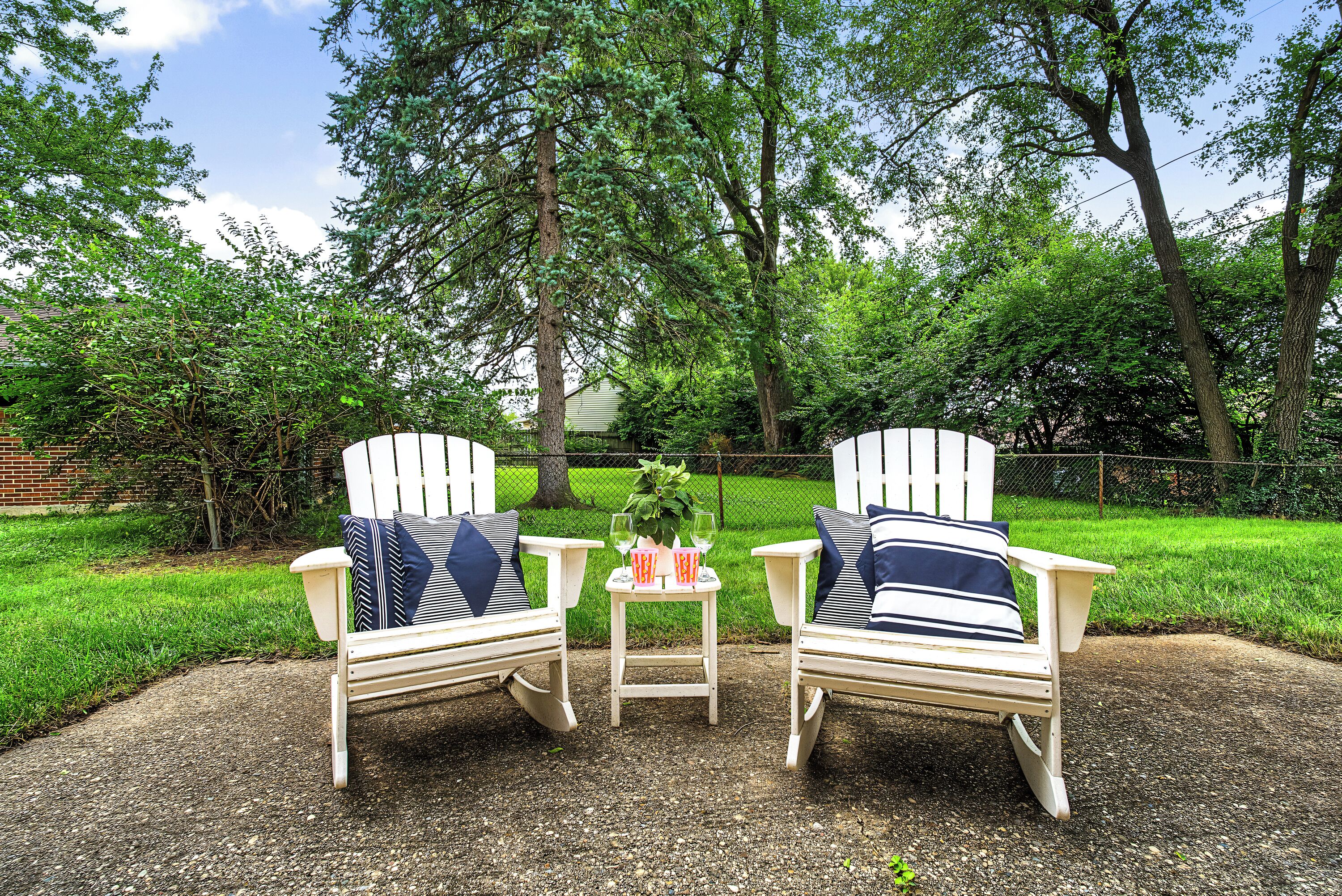 Two Adirondack chairs with striped cushions, a small table with a glass of wine and a pink cup, set on a gravel surface with a grassy lawn and trees in the background.