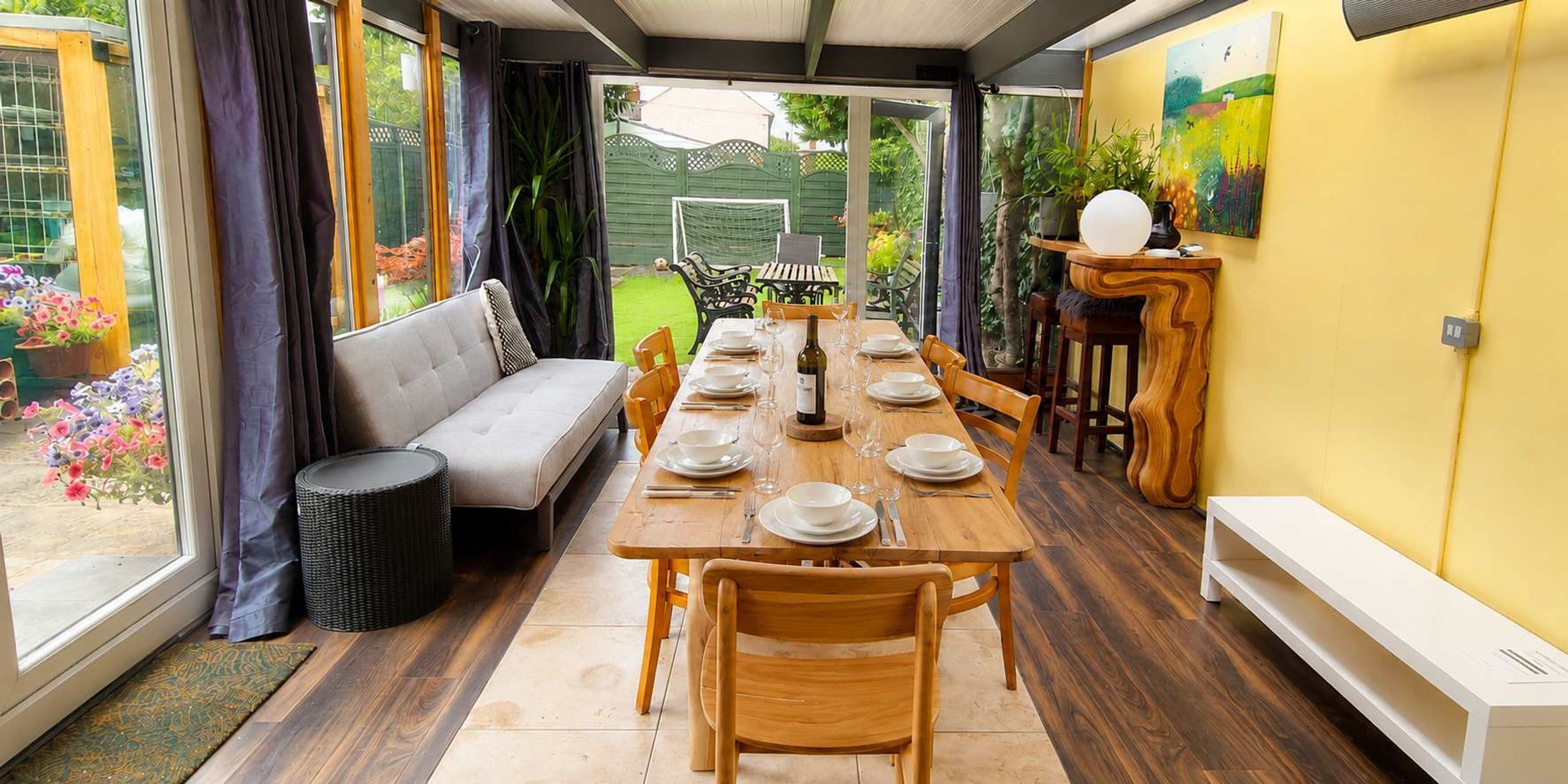Un coin repas avec une longue table dressée pour six personnes, entourée de chaises en bois, d’un canapé et d’une petite table d’appoint avec une lampe.