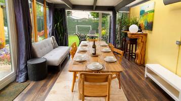 A dining area with a long table set for six, surrounded by wooden chairs, a sofa, and a small side table with a lamp.