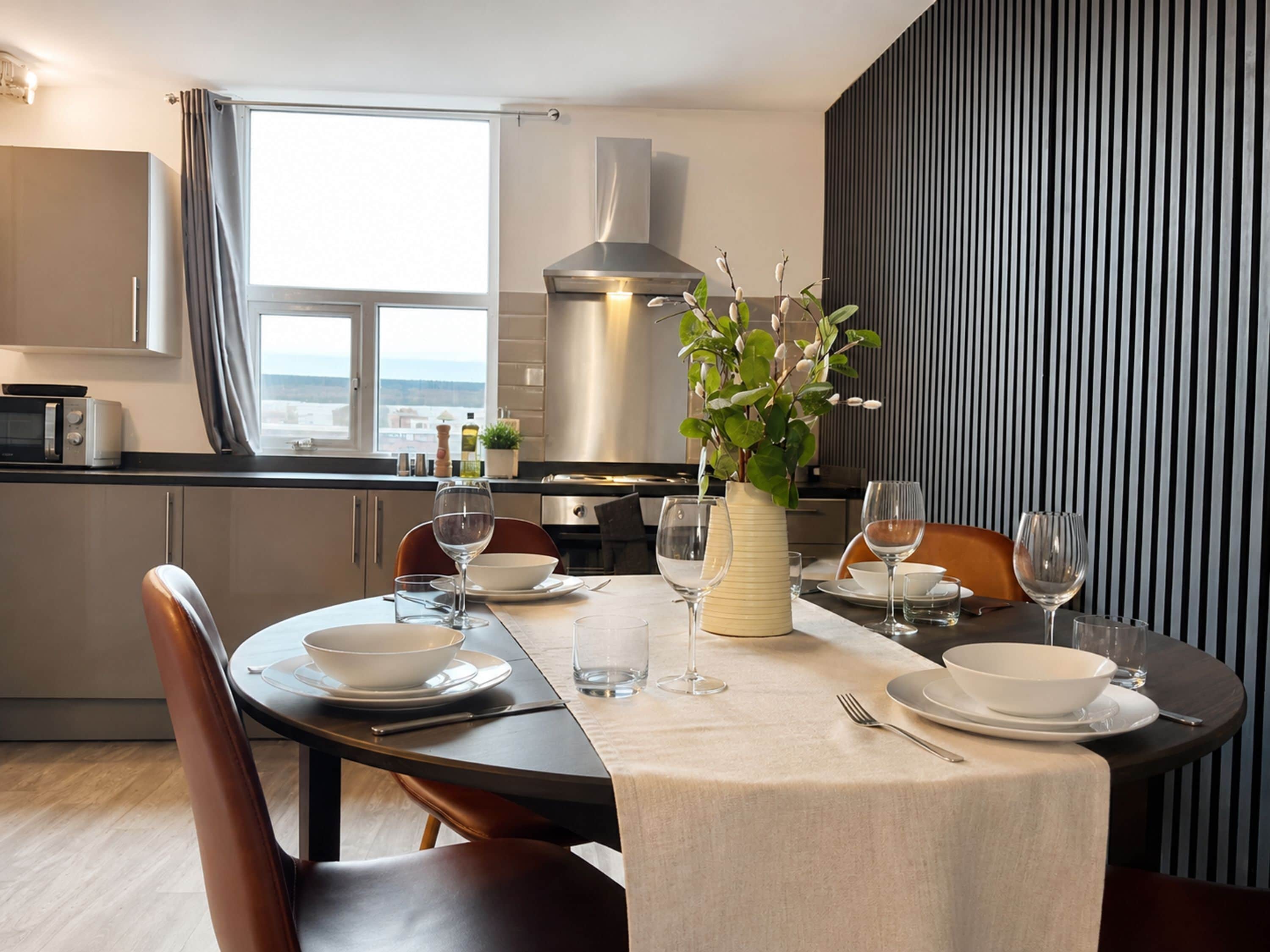 A modern dining area with a table set for two, a vase with flowers, and a view of the sea through the window.