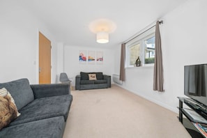 A modern living room with a grey sofa, a small side table, a grey armchair, a flat-screen TV, and abstract wall art.