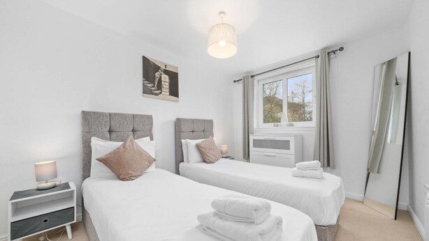 A hotel room with two beds, a bedside table with a lamp, a window with curtains, and a picture on the wall. - Sleek 2BR Edinburgh Near Murrayfield Sleeps 4 (Edinburgh)