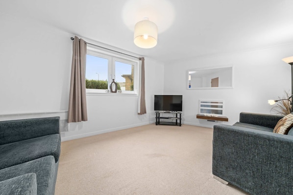 A modern living room with a flat-screen TV, grey sofas, a large window with curtains, and a ceiling light fixture.