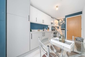 A modern kitchen with white cabinets, a blue accent wall, and a glass dining table with a vase.